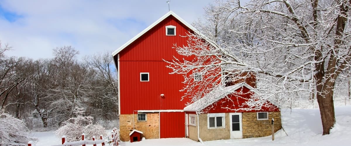A red barn with a stone base, surrounded by snow-covered trees and a wooden fence.