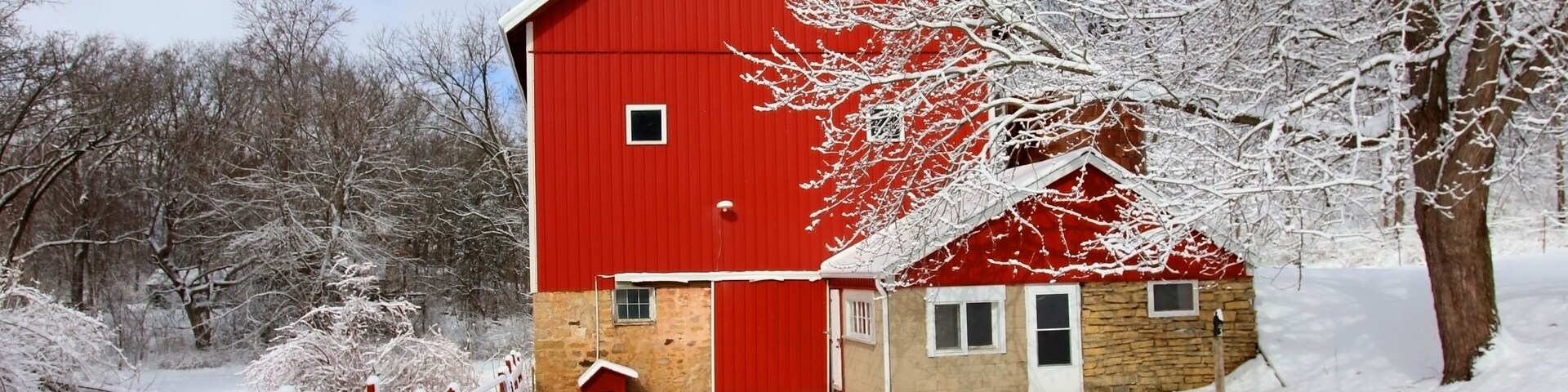 A red barn with a stone base, surrounded by snow-covered trees and a wooden fence.