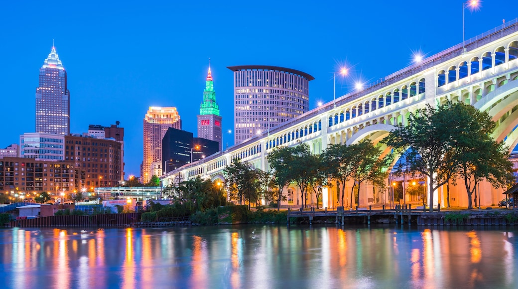 A city skyline at dusk with illuminated buildings, a river reflecting lights, and a bridge with arches.
