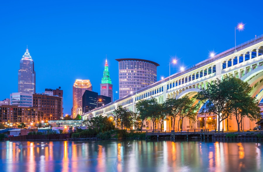 A city skyline at dusk with illuminated buildings, a river reflecting lights, and a bridge with arches.