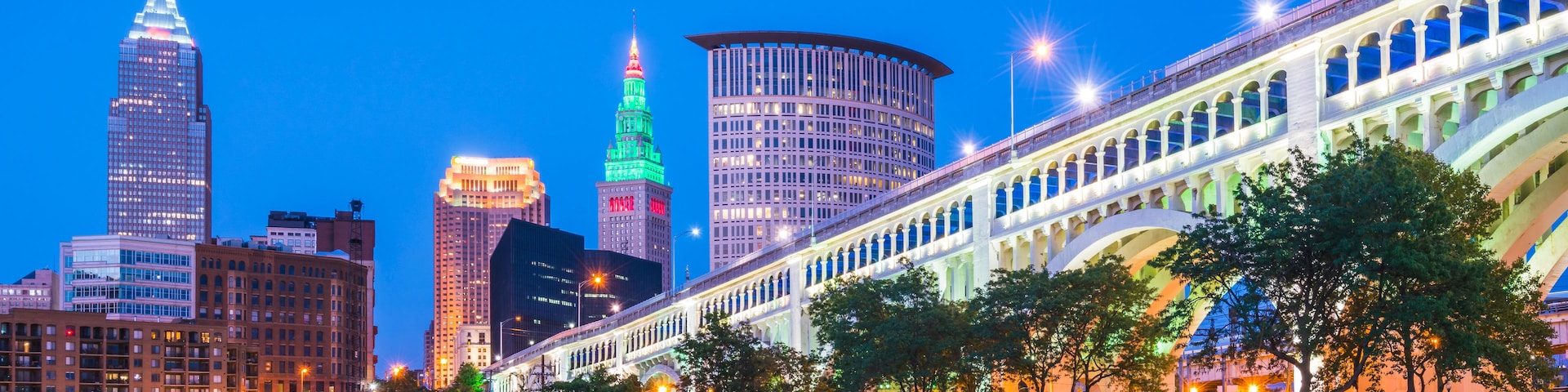 Linha do horizonte urbano ao entardecer, com edifícios iluminados, um rio refletindo luzes e uma ponte com arcos.