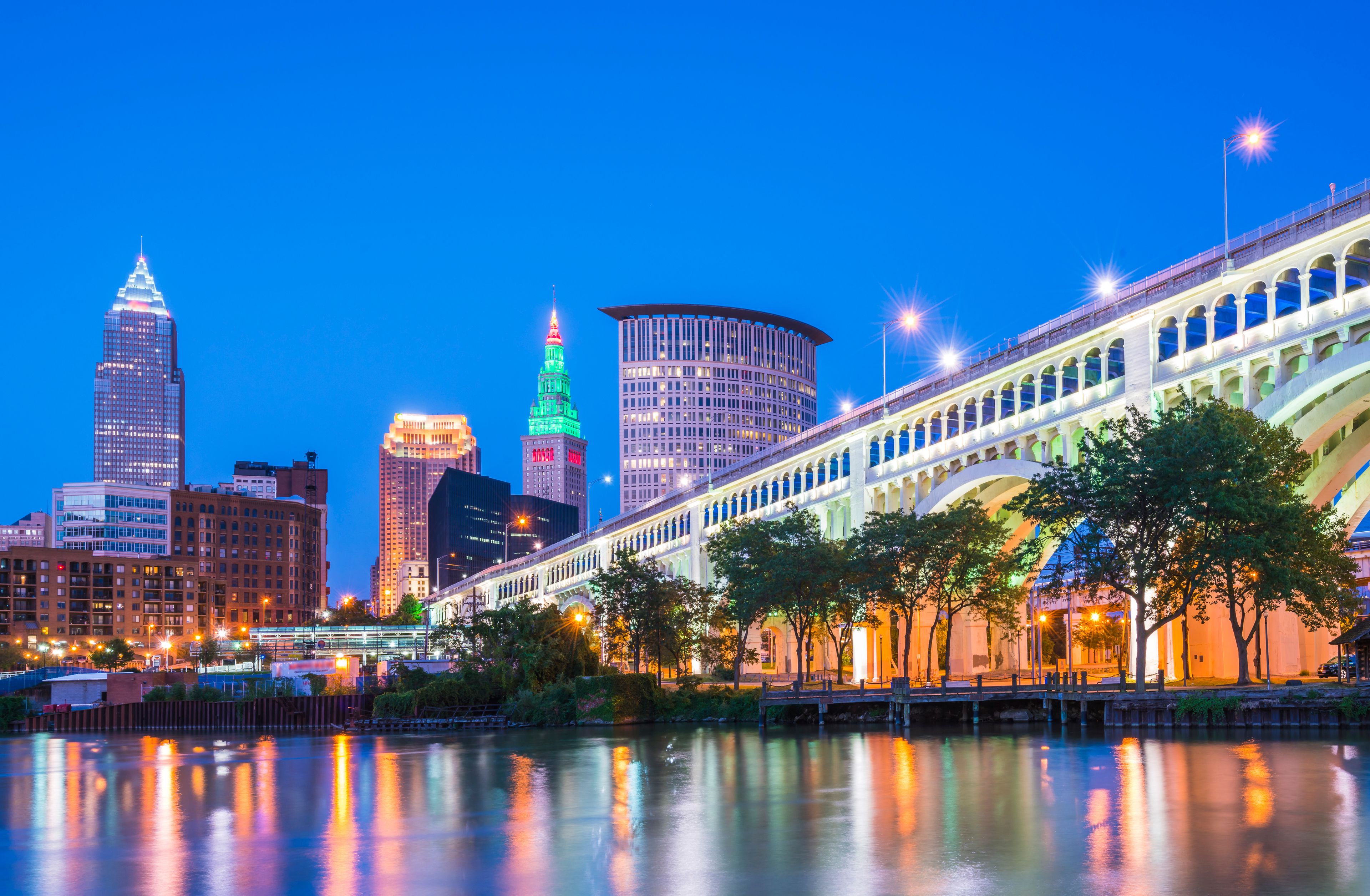 A city skyline at dusk with illuminated buildings, a river reflecting lights, and a bridge with arches.