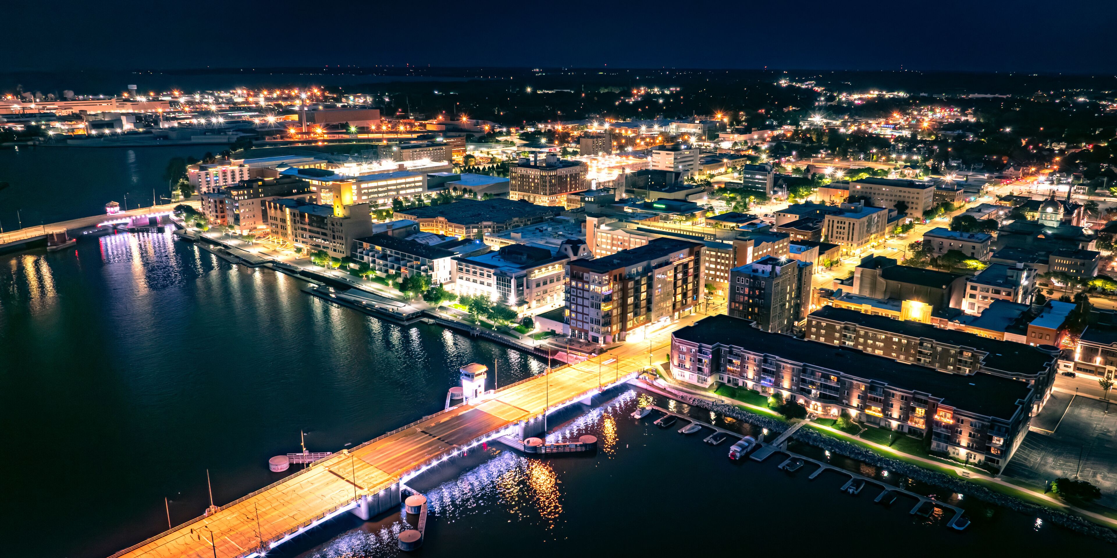 A nighttime cityscape with a waterfront, illuminated buildings, and a bridge.