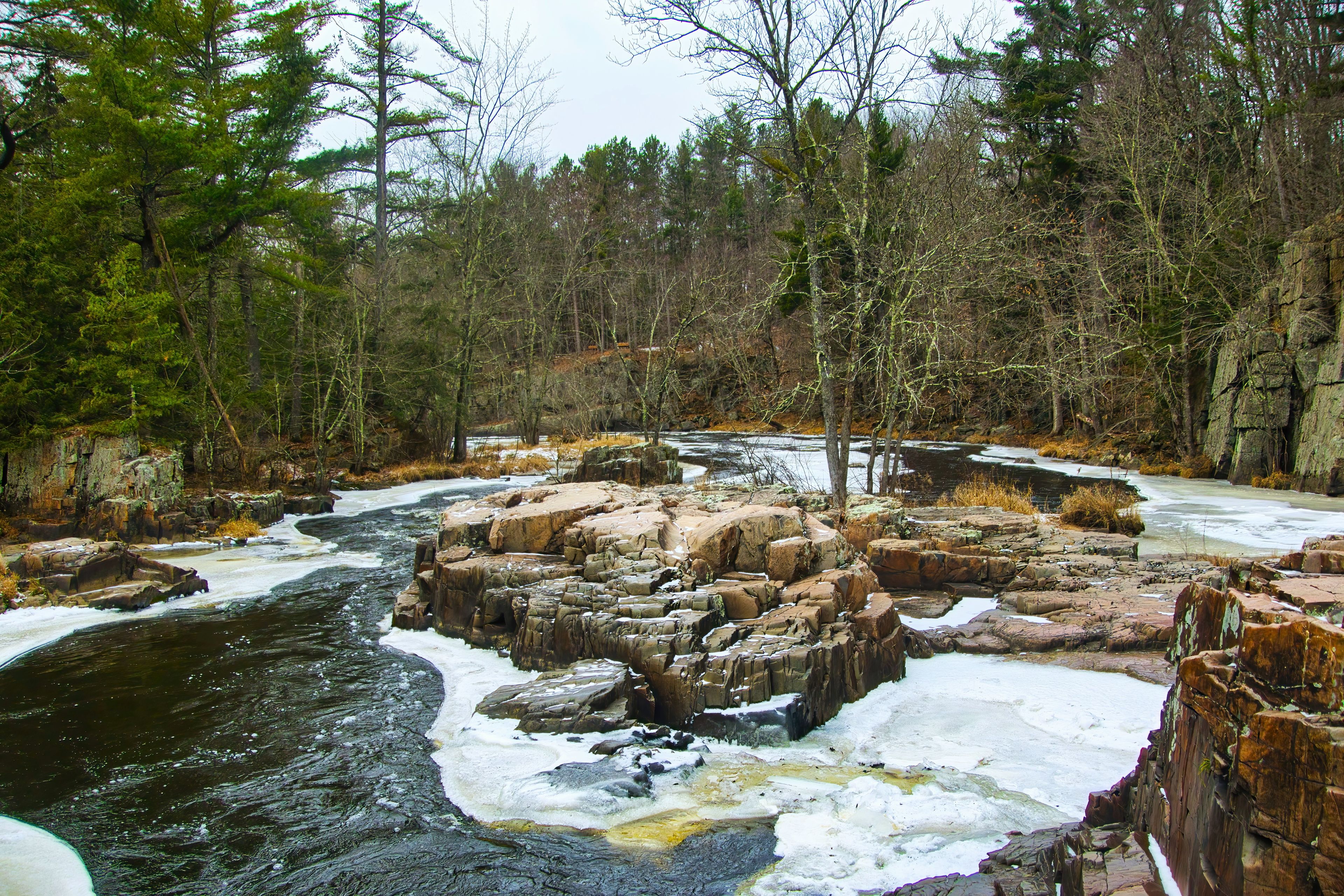 Ein Fluss fließt durch einen Wald mit felsigen Vorsprüngen und Schneeflecken.