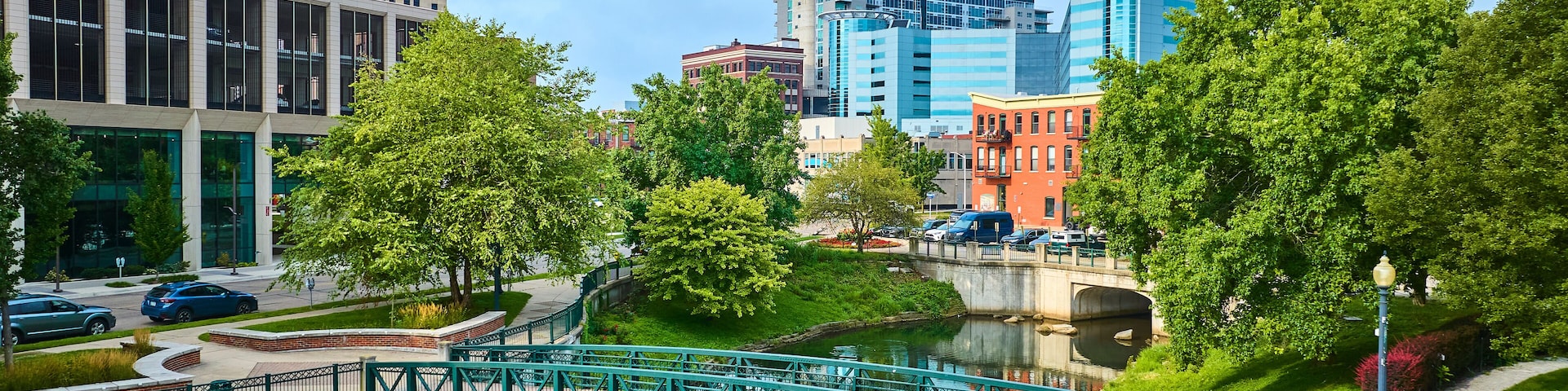 Uma ponte verde sobre um canal, rodeada por vegetação exuberante e edifícios modernos.