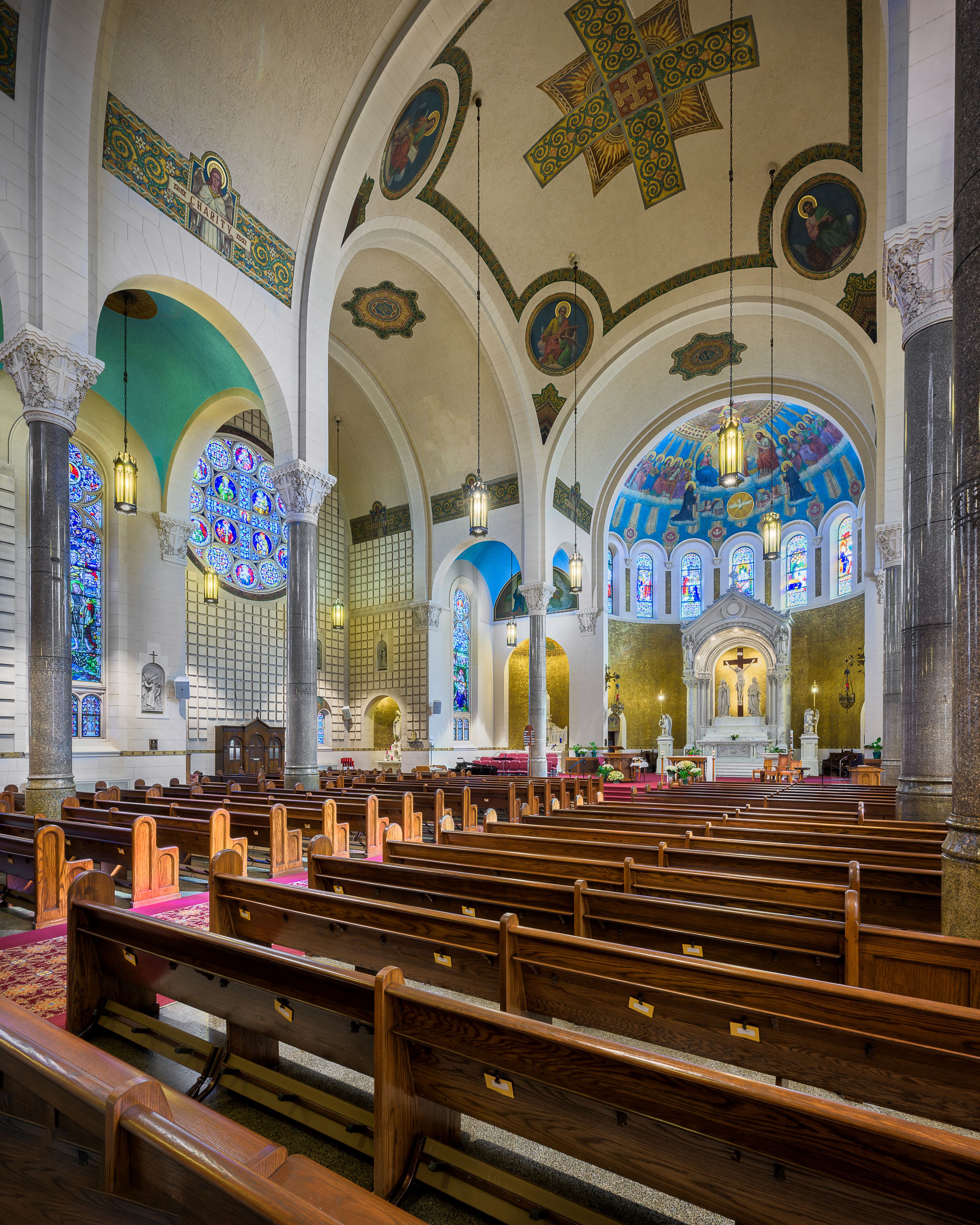 A church interior with wooden pews, stained glass windows, and ornate ceiling decorations.