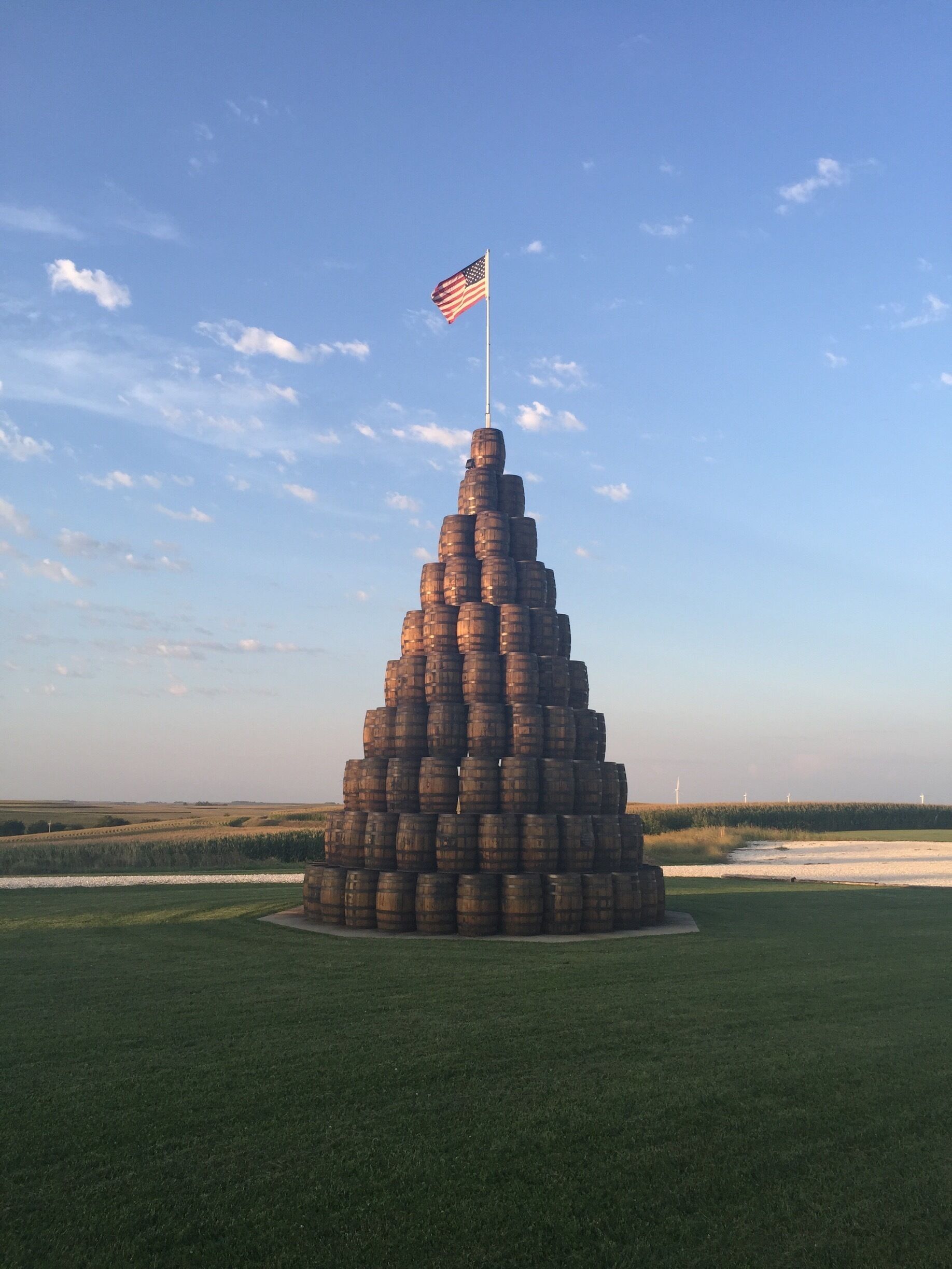 A pyramid of barrels with an American flag on top, set in a grassy field with wind turbines in the background.