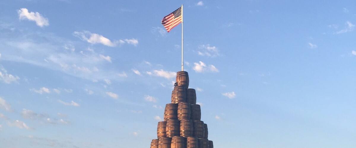 A pyramid of barrels with an American flag on top, set in a grassy field with wind turbines in the background.