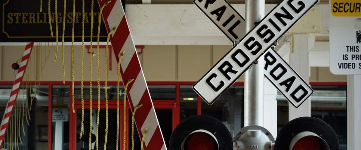A railroad crossing sign with a red and white striped gate, a red light, and a security sign.