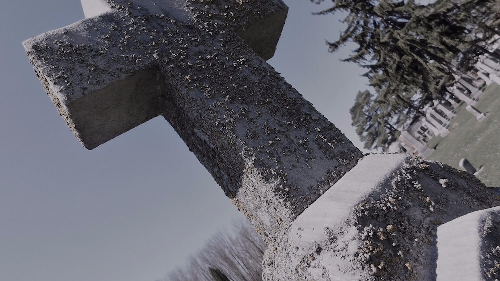 Une croix de pierre recouverte de neige, avec un cimetière en arrière-plan.