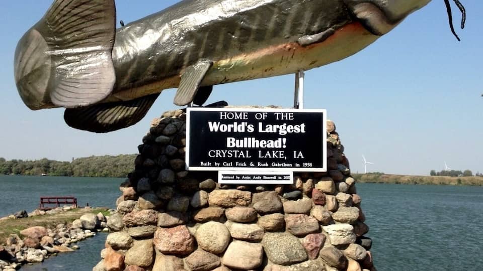 A large fish sculpture on a stone pedestal with a sign stating it's the home of the world's largest bullhead fish in Crystal Lake, IA.