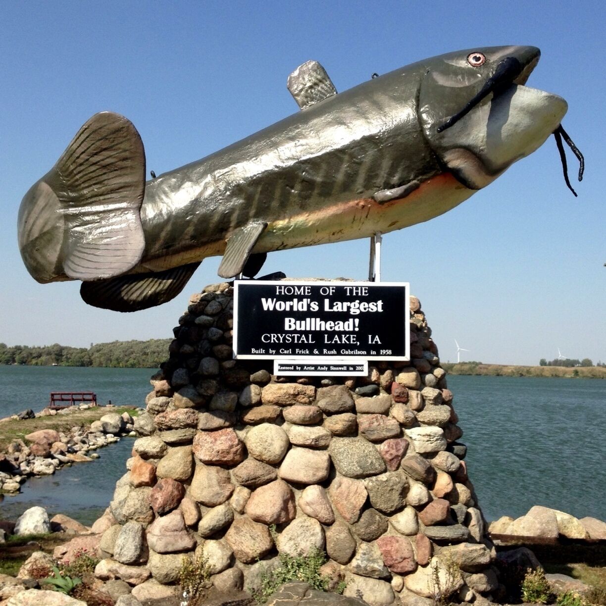 A large fish sculpture on a stone pedestal with a sign stating it's the home of the world's largest bullhead fish in Crystal Lake, IA.