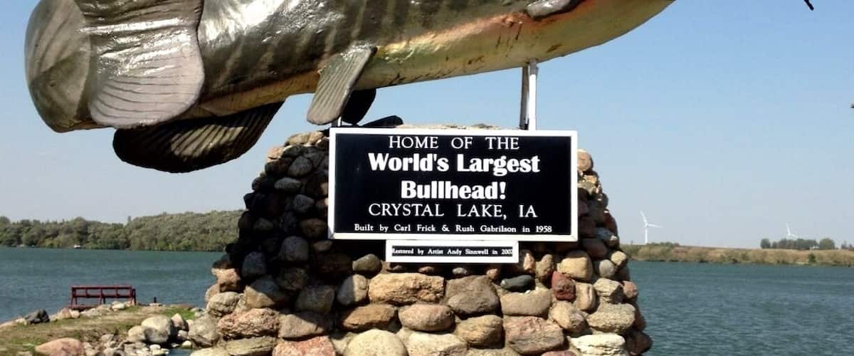 A large fish sculpture on a stone pedestal with a sign stating it's the home of the world's largest bullhead fish in Crystal Lake, IA.
