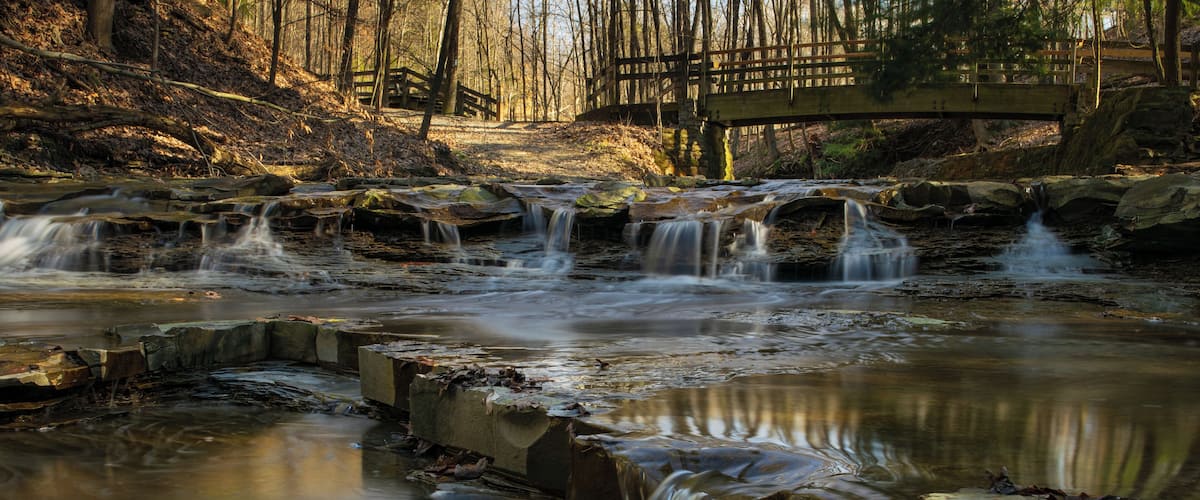 A waterfall in a forested area with a wooden bridge in the background.