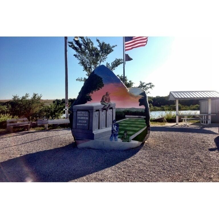 A sculpture of a man sitting on a rock with a sunset in the background, an American flag, and a small building in the foreground.