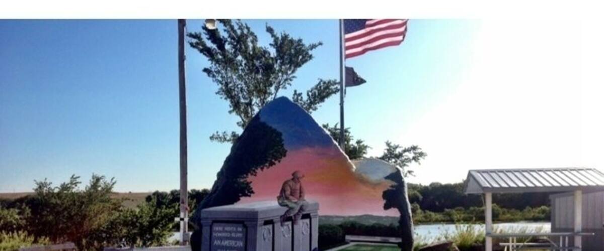 A sculpture of a man sitting on a rock with a sunset in the background, an American flag, and a small building in the foreground.