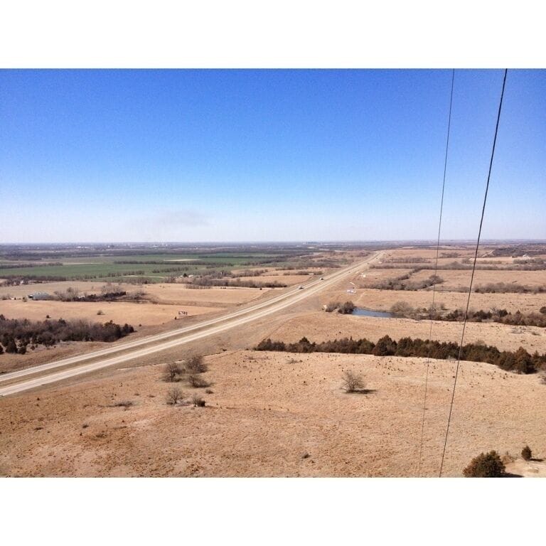 A long, straight road through a dry, open landscape with sparse vegetation and distant fields.