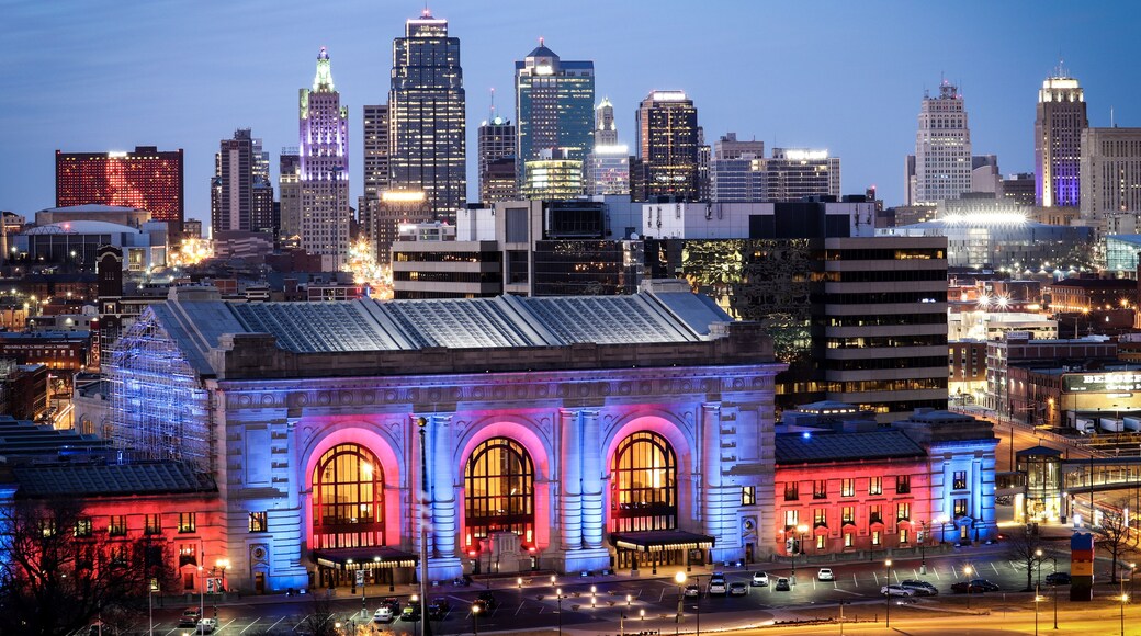 A city skyline at dusk with illuminated buildings and a prominent historic building with arched windows.