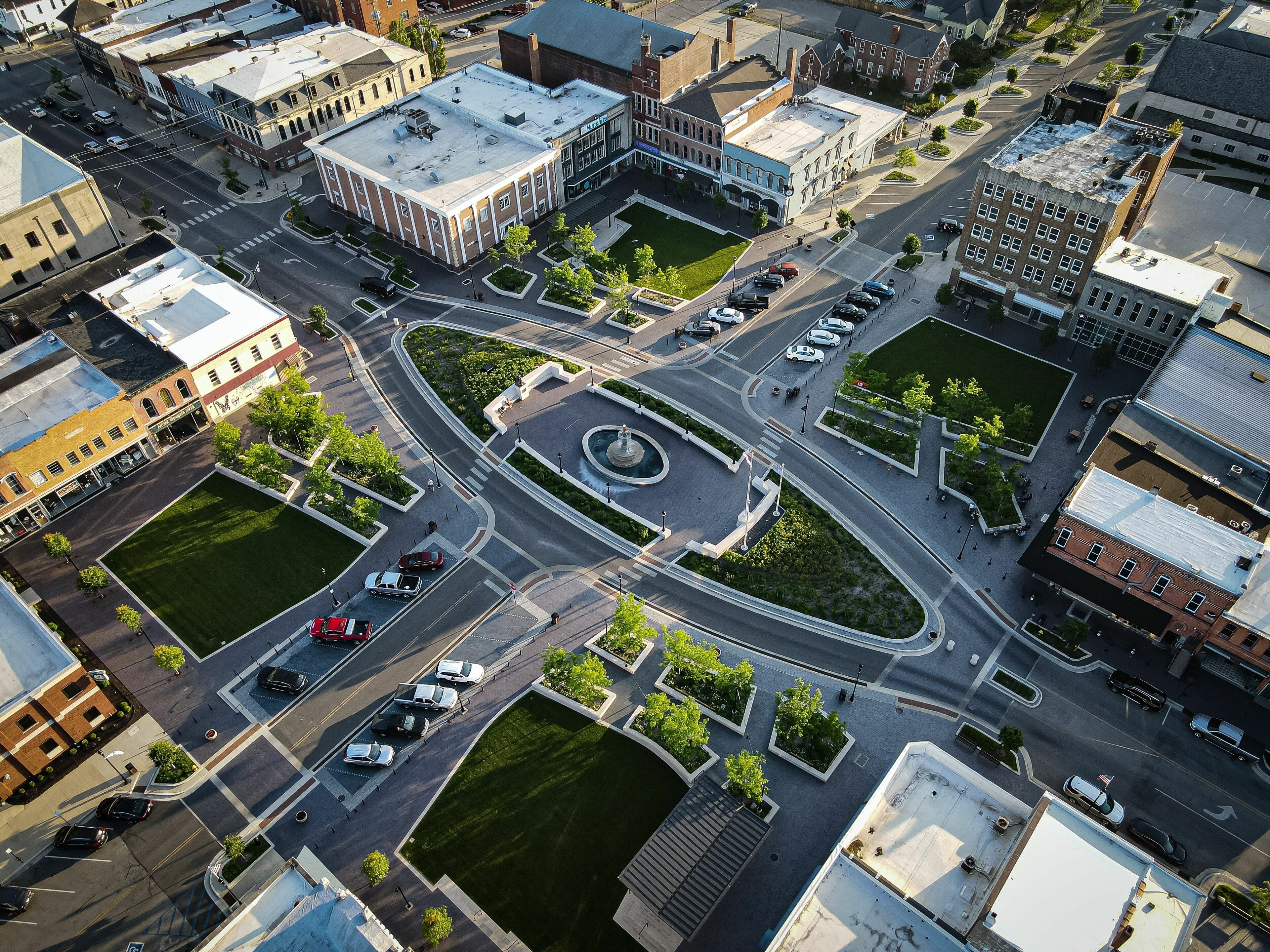 An aerial view of a city intersection with a roundabout, surrounding buildings, and a central fountain.