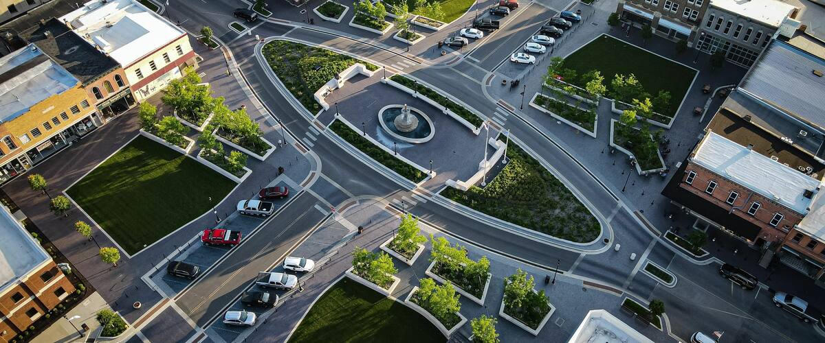 An aerial view of a city intersection with a roundabout, surrounding buildings, and a central fountain.