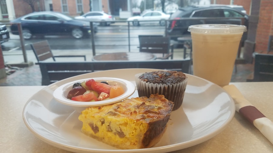 A view from inside looking out to the patio and street beyond.
The food on the plate is a slice of their Sausage & Bacon Quiche, bowl of fresh fruit, and the Chocolate Reeses Muffin. Also photographed is the medium size of an Iced Chai Tea Latte.