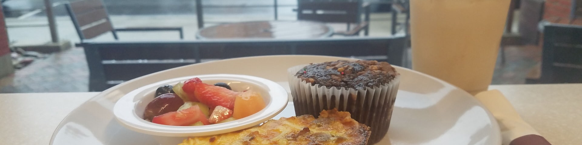 A view from inside looking out to the patio and street beyond.
The food on the plate is a slice of their Sausage & Bacon Quiche, bowl of fresh fruit, and the Chocolate Reeses Muffin. Also photographed is the medium size of an Iced Chai Tea Latte.