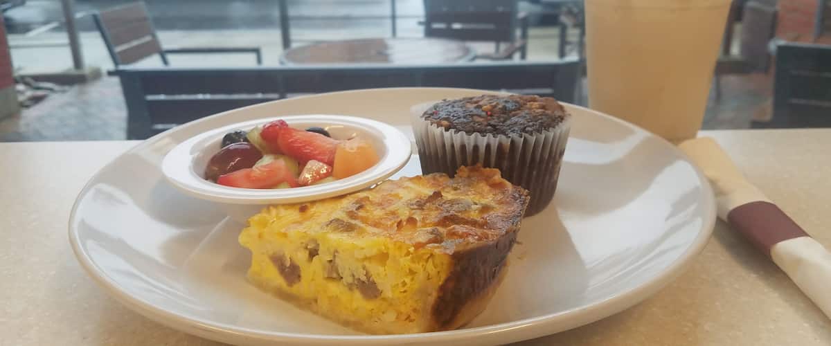 A plate with a slice of quiche, a muffin, and a cup of iced coffee on a table.