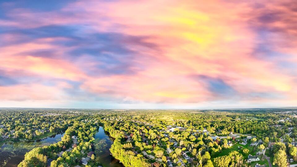 A panoramic view of a town with a river running through it, surrounded by dense greenery and a colorful sky.