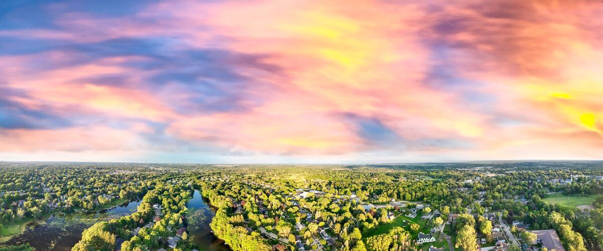 A panoramic view of a town with a river running through it, surrounded by dense greenery and a colorful sky.