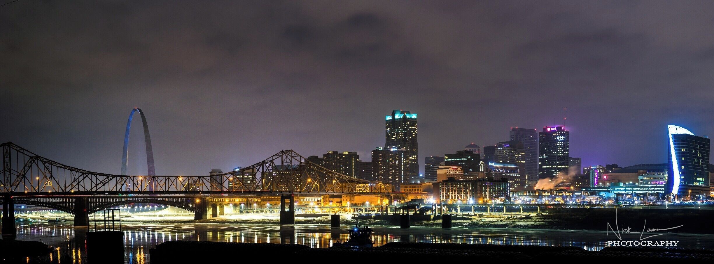 A city skyline at night with illuminated buildings, a bridge, and a river reflecting the lights.