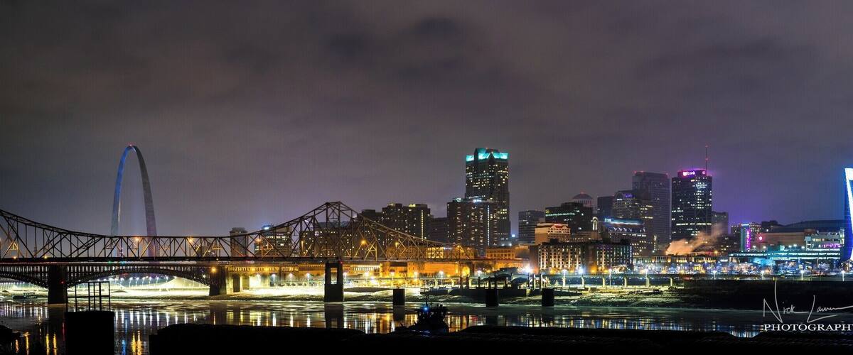 A city skyline at night with illuminated buildings, a bridge, and a river reflecting the lights.