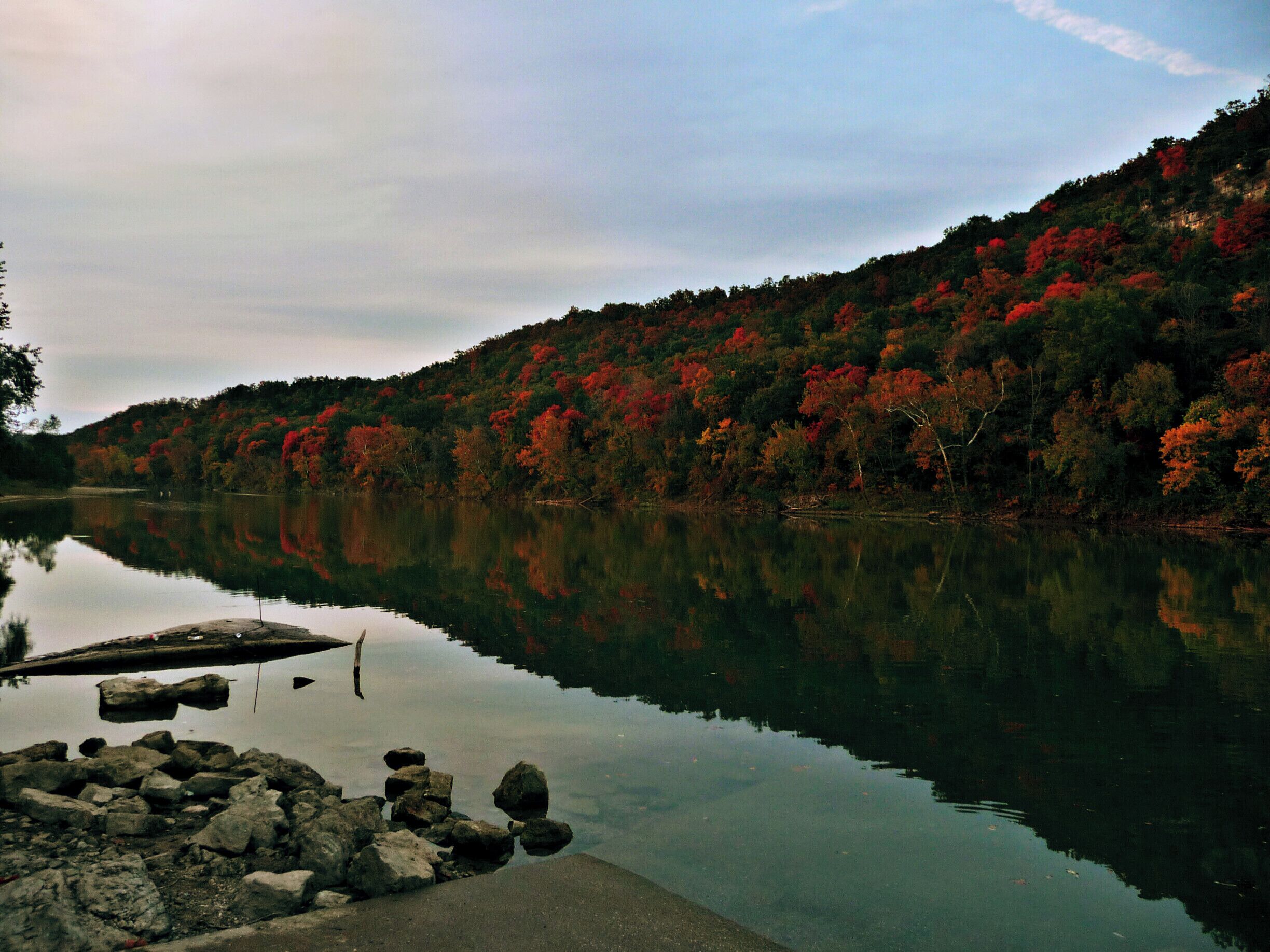 A calm lake with a dock and rocks in the foreground, surrounded by a forest with autumn foliage.