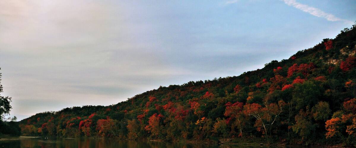 A calm lake with a dock and rocks in the foreground, surrounded by a forest with autumn foliage.