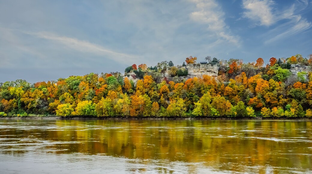 A river reflecting autumn foliage, a rocky cliff, and a partly cloudy sky.