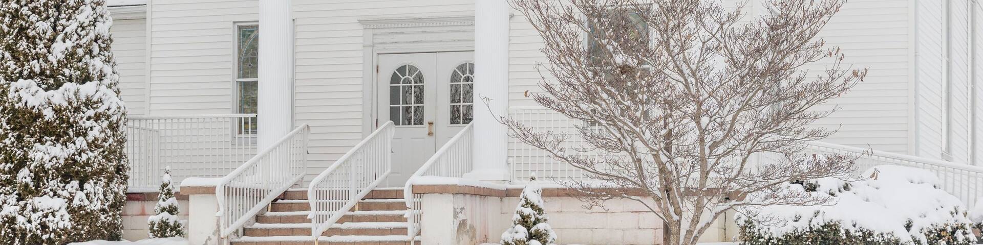 A white church with a steeple and a cross on top, surrounded by snow-covered trees.