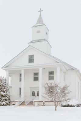 A white church with a steeple and a cross on top, surrounded by snow-covered trees.