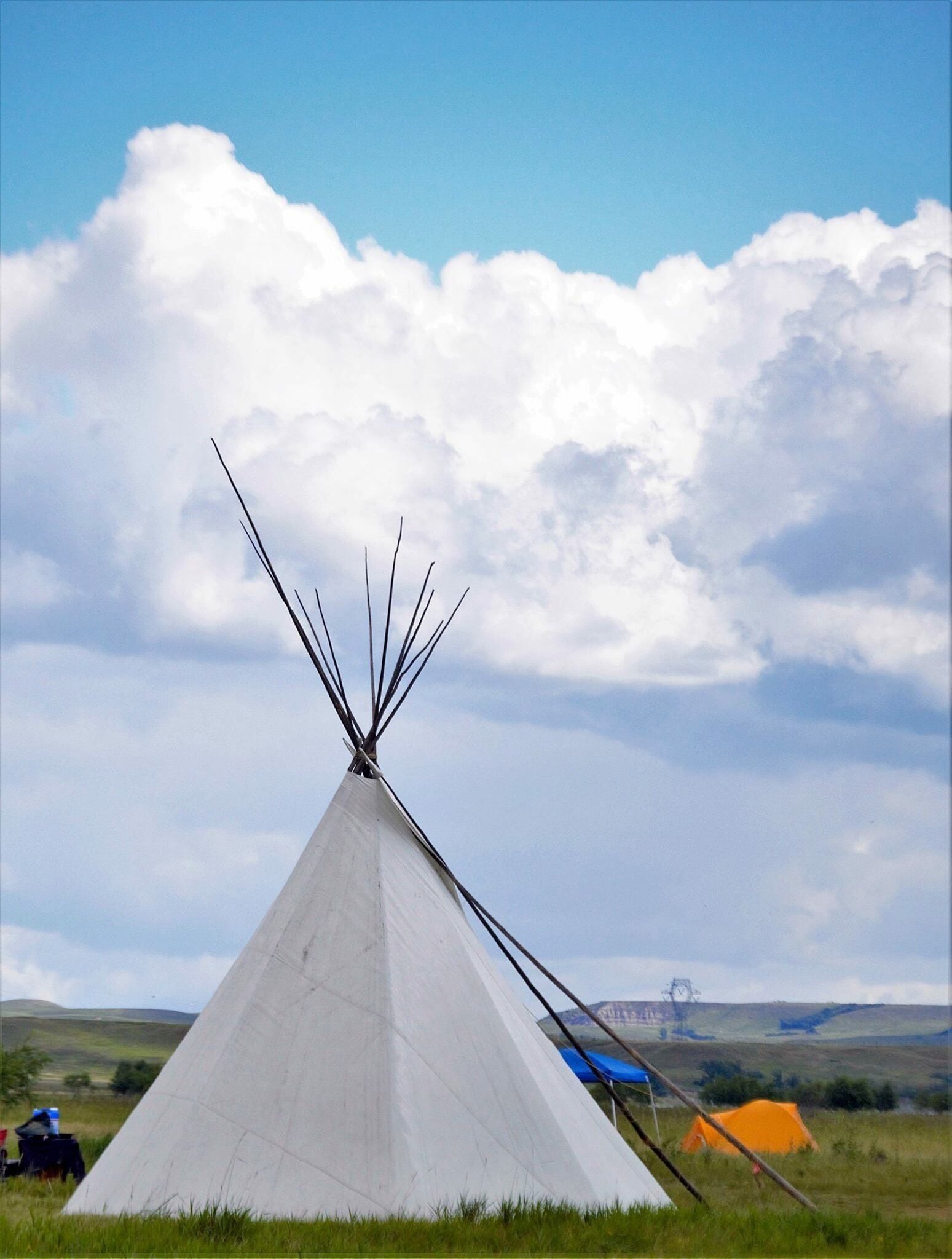 A white tipi with a pointed top, set in a grassy field with a blue sky and clouds.