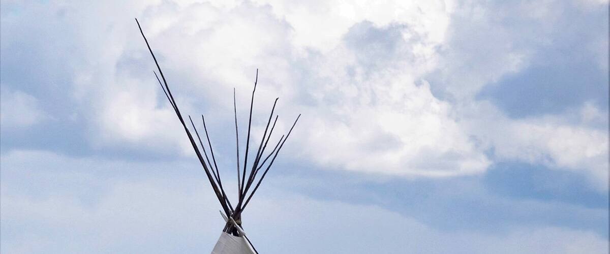 A white tipi with a pointed top, set in a grassy field with a blue sky and clouds.