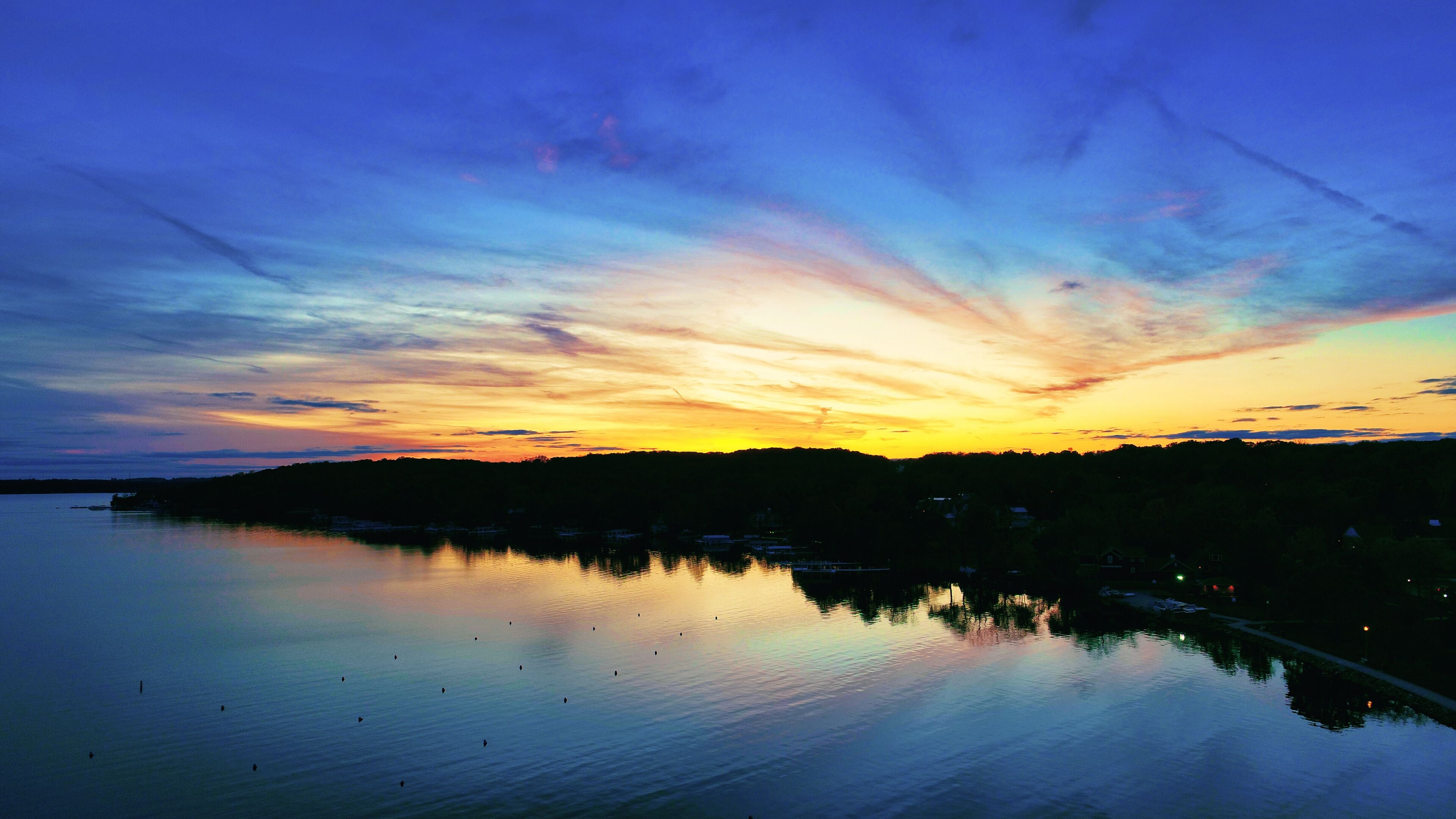 A sunset over a calm lake with a silhouette of a forested hill and a row of houses along the shore.