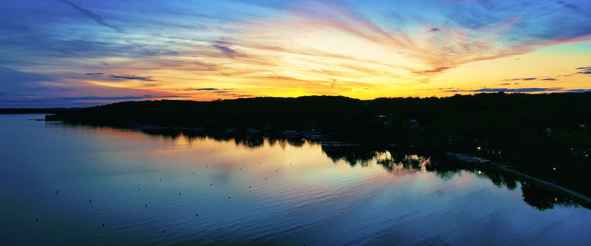 A sunset over a calm lake with a silhouette of a forested hill and a row of houses along the shore.