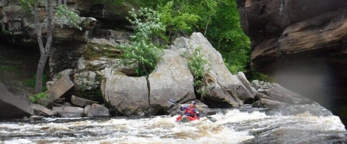A kayaker navigating a turbulent river surrounded by rocky terrain and dense greenery.