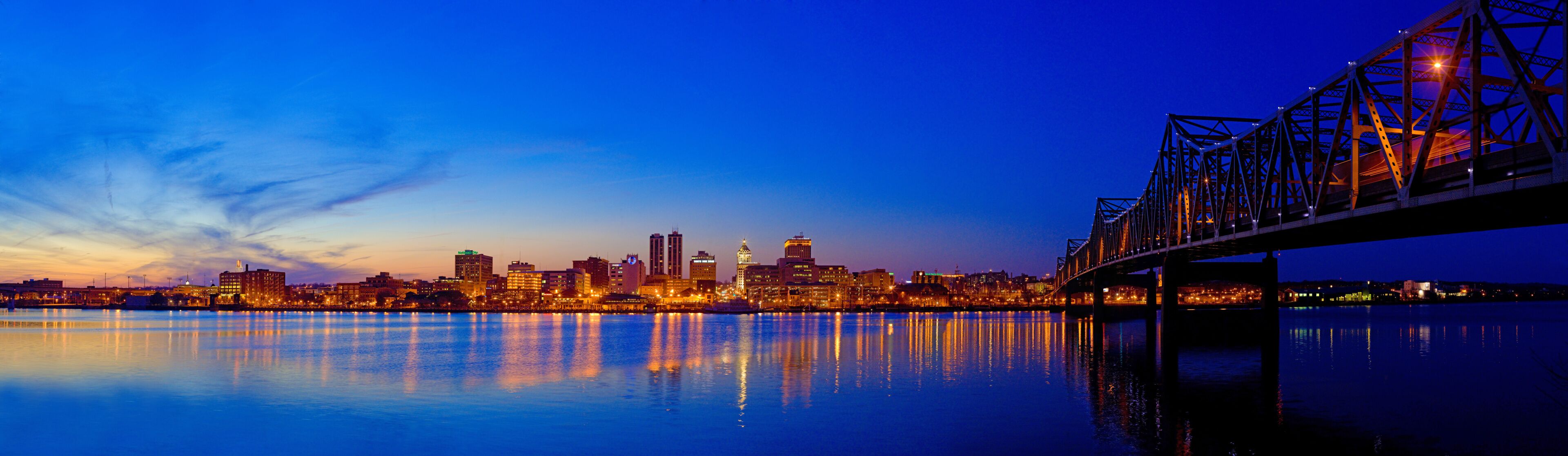 A city skyline at dusk with a bridge reflecting on the water.