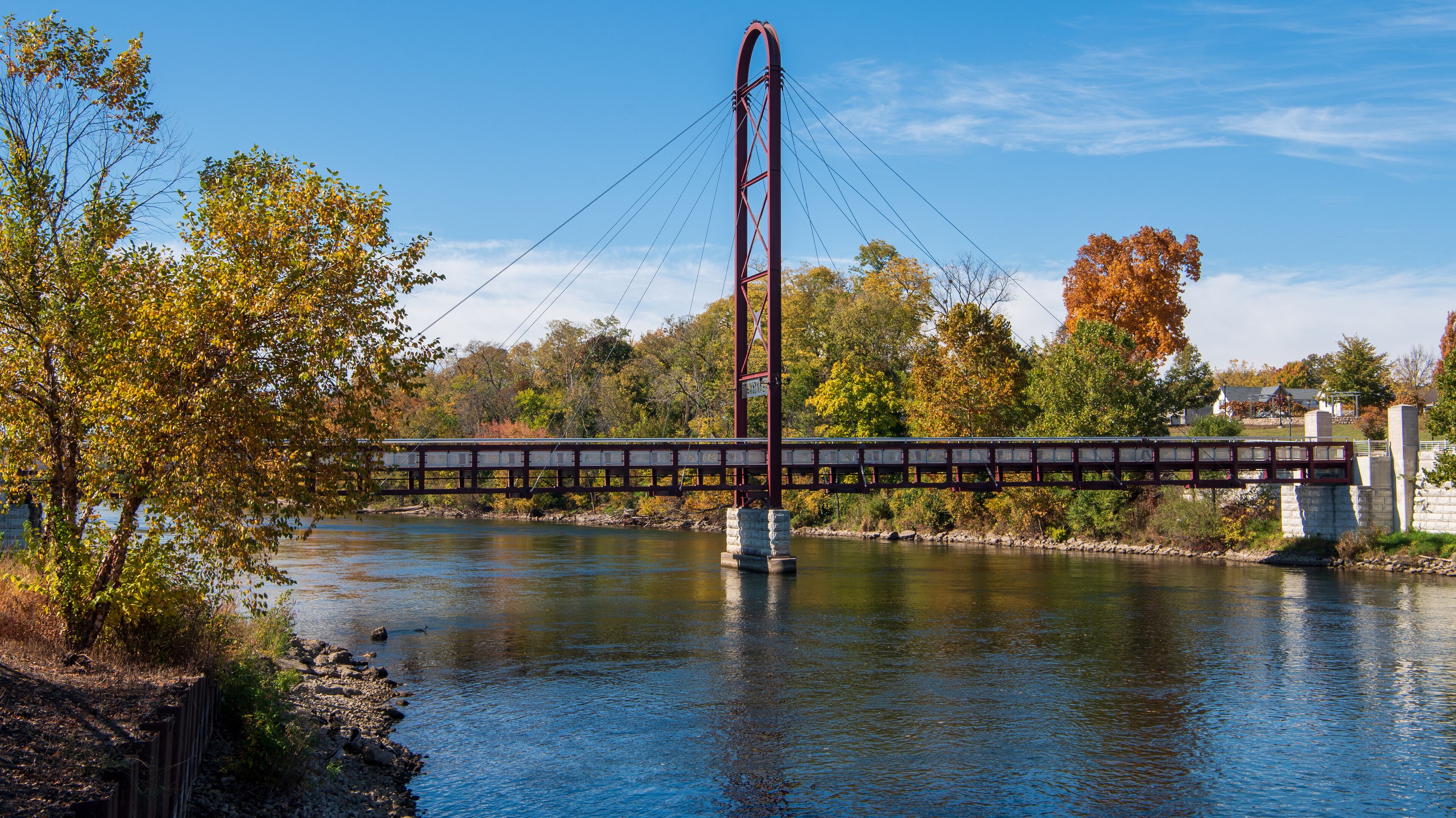 A suspension bridge over a river with autumn foliage and houses in the background.