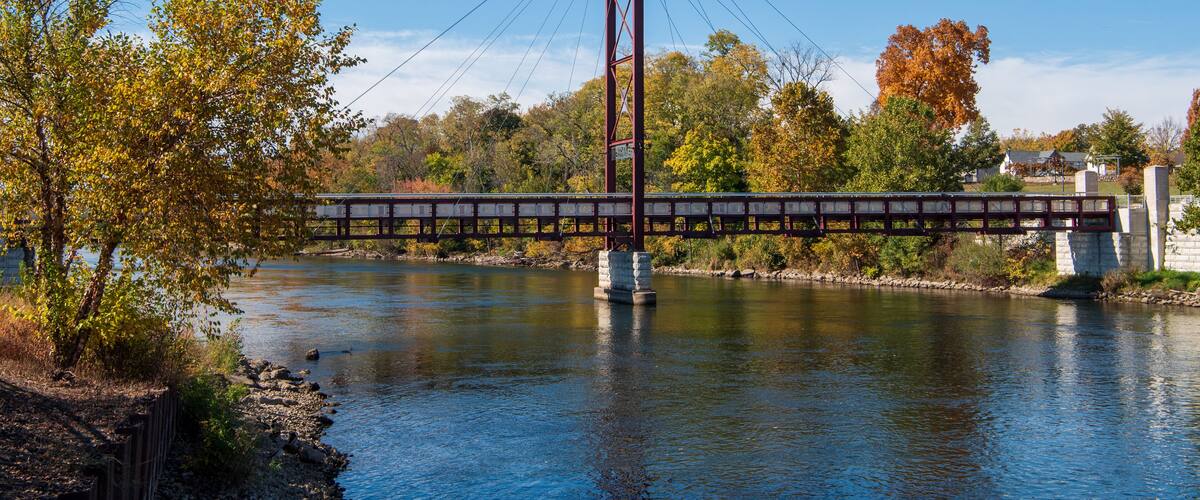 A suspension bridge over a river with autumn foliage and houses in the background.