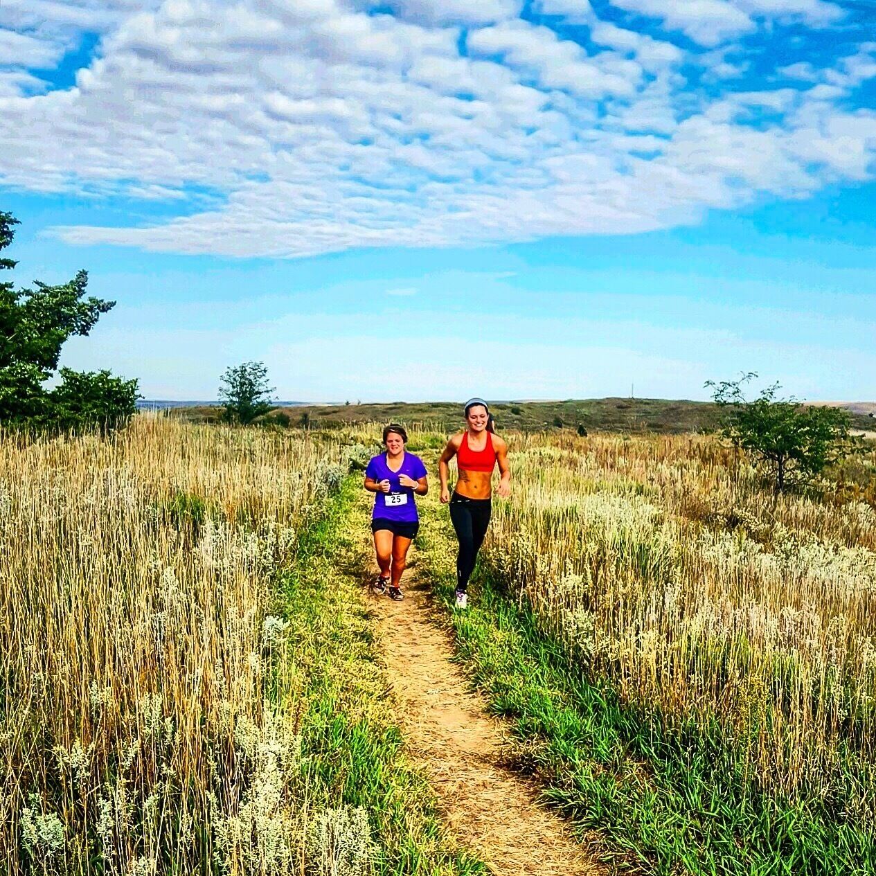 Two people running on a dirt path through a field with tall grass and a clear sky.