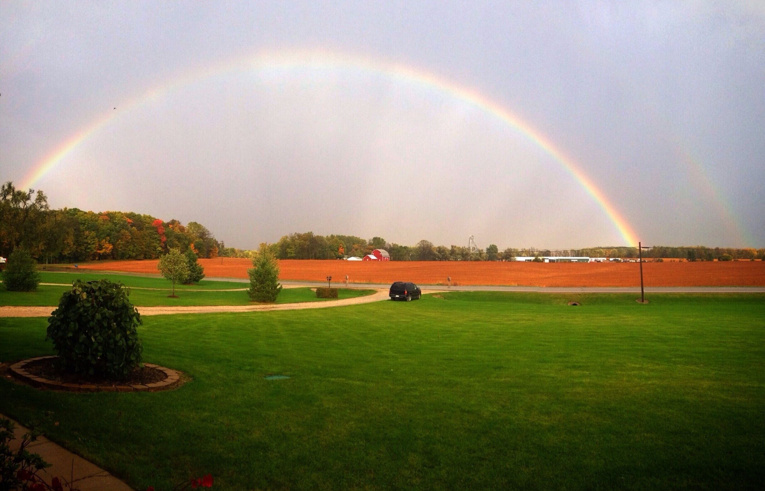 A rainbow over a field with a car and a red barn.
