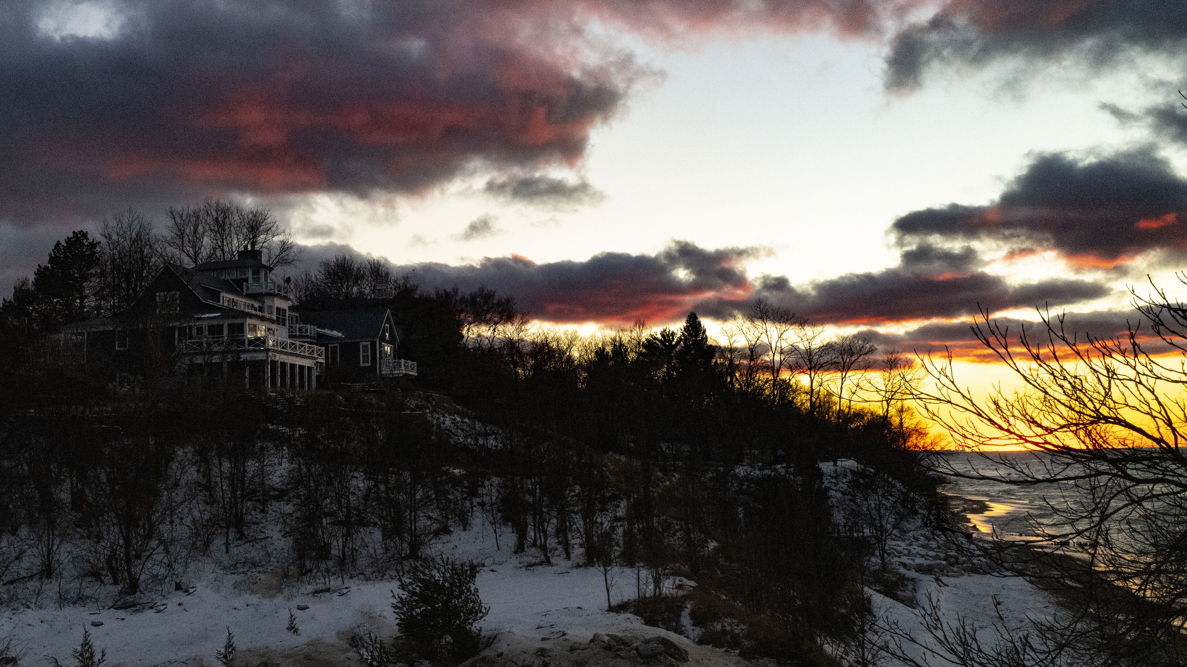 A snow-covered landscape with a house on a cliff overlooking a sunset.