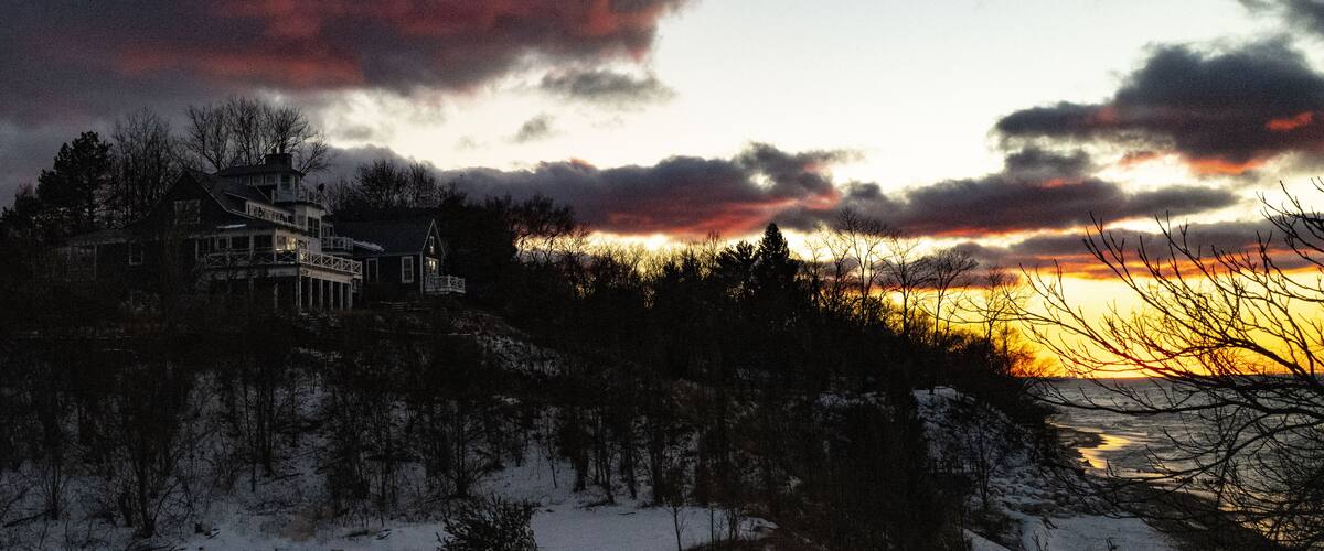 A snow-covered landscape with a house on a cliff overlooking a sunset.