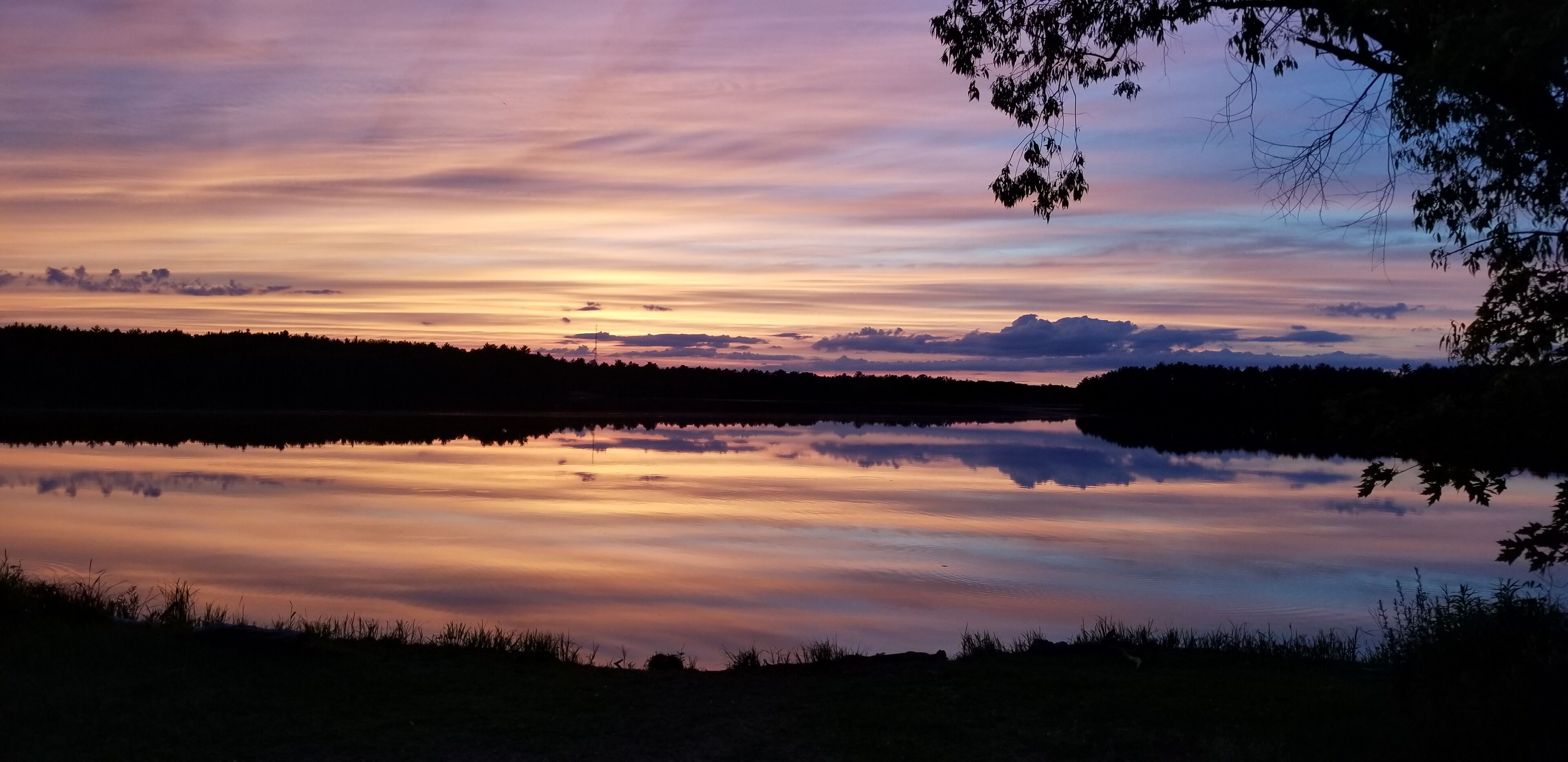 A sunset over a calm lake with reflections of the sky, surrounded by trees and a distant mountain range.