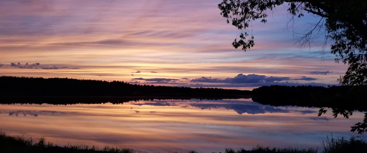 A sunset over a calm lake with reflections of the sky, surrounded by trees and a distant mountain range.